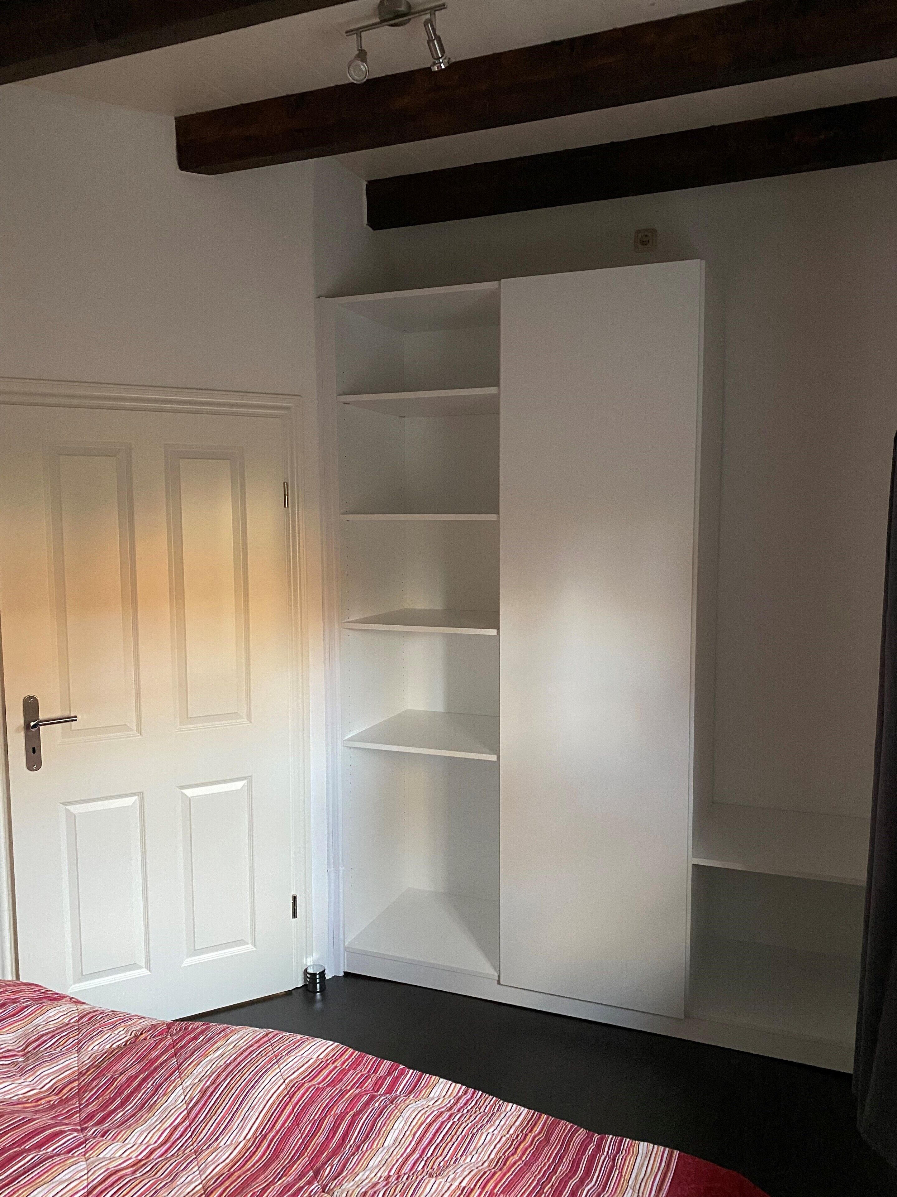 Una habitación con una puerta blanca, una cama con colcha a rayas y una estantería empotrada blanca con varios compartimentos.