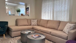 En stue med en beige hjørnesofa, to runde sofaborde, en blød måtte og en grøn lænestol.