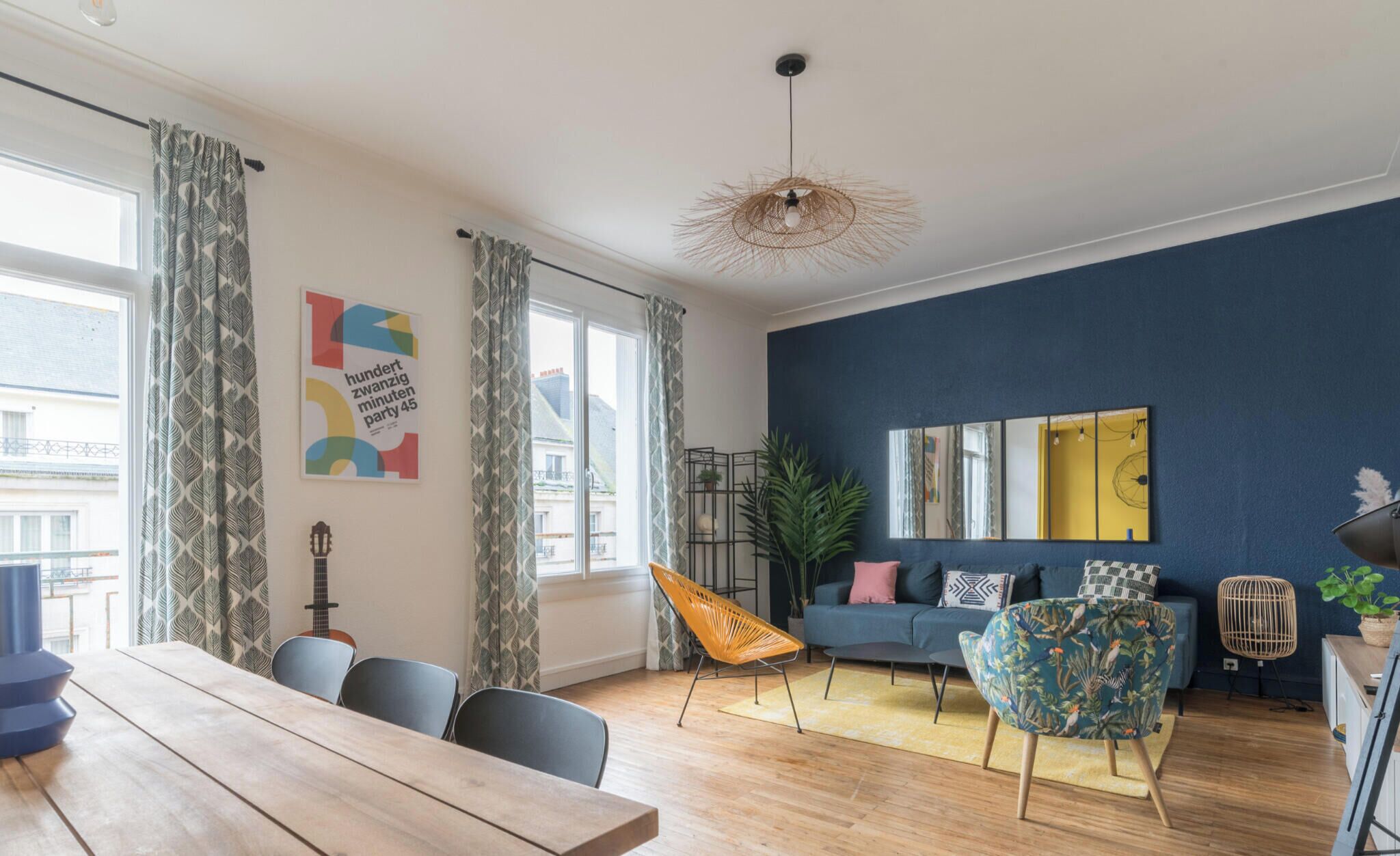 A modern living room with a wooden dining table, a sofa, and a chair. There is a guitar leaning against the wall, a plant, and a mirror on the wall.