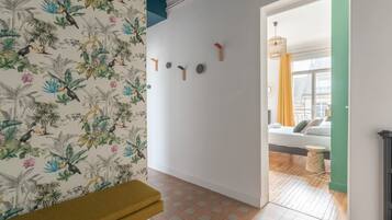 Une pièce avec un papier peint à motifs, un banc, une horloge murale et une vue sur une autre pièce où se trouvent un lit et une fenêtre.