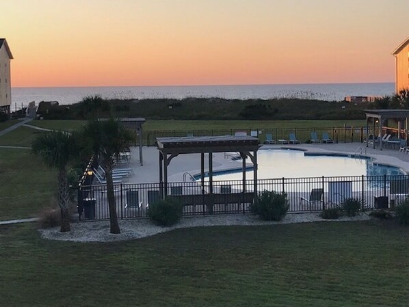 A pool area with a gazebo, a fence, and a view of the ocean at sunset.