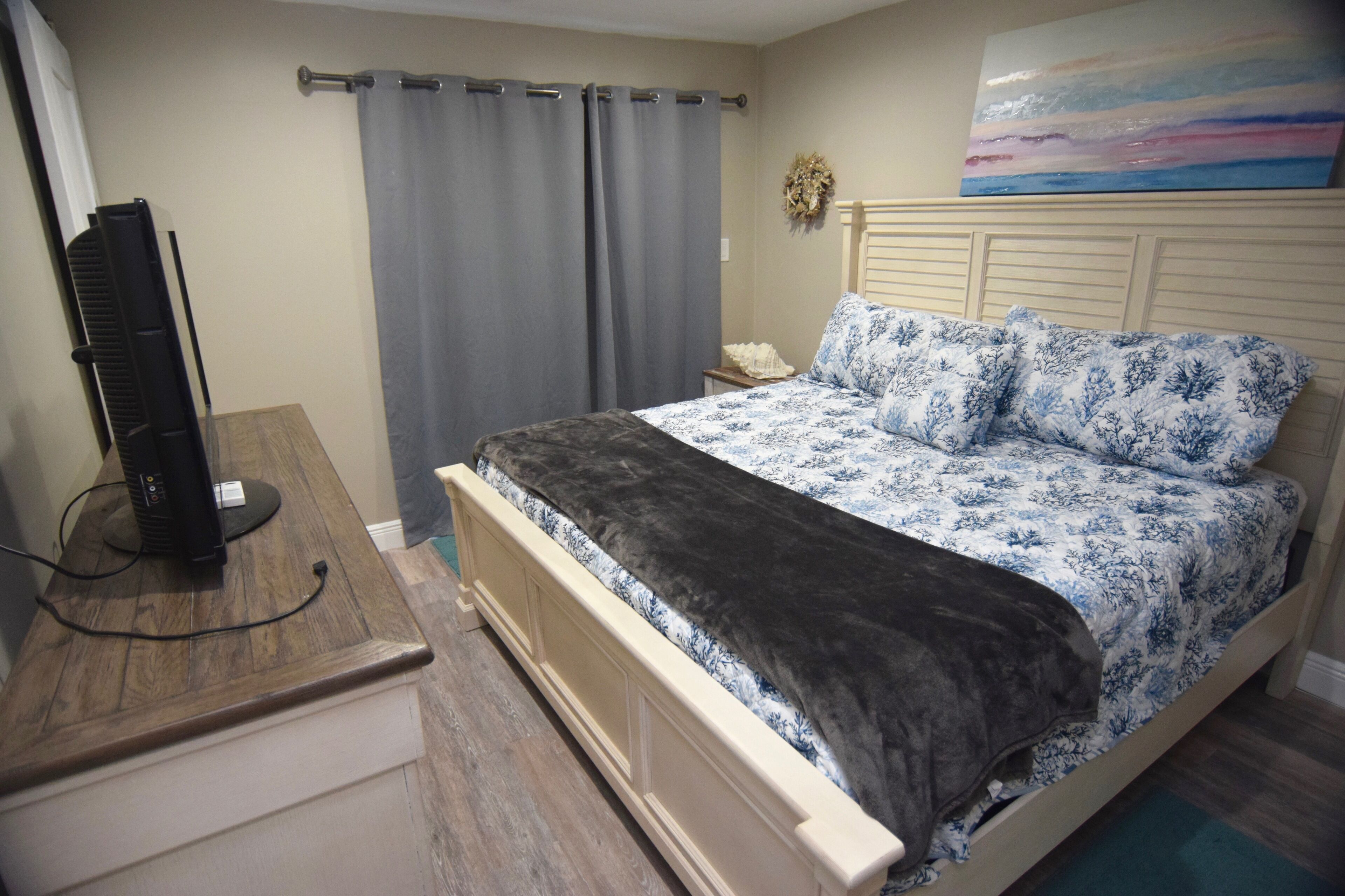 A bedroom with a bed, a desk with a computer, a nightstand, a painting on the wall, and a window with curtains.