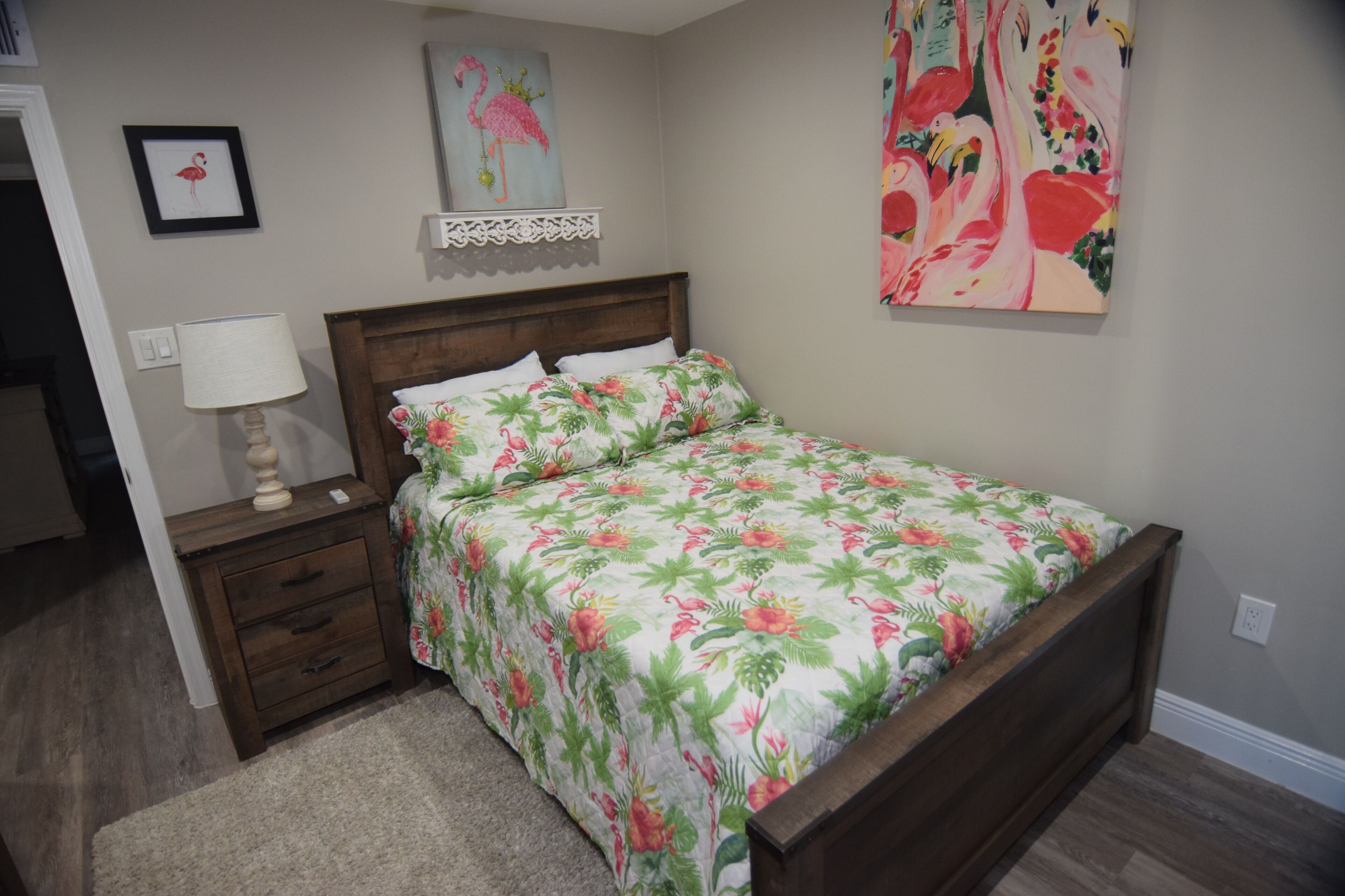 Une chambre avec un lit en bois, une literie à motifs floraux, une table de chevet avec une lampe et un mur orné d’un tableau représentant un flamant rose.