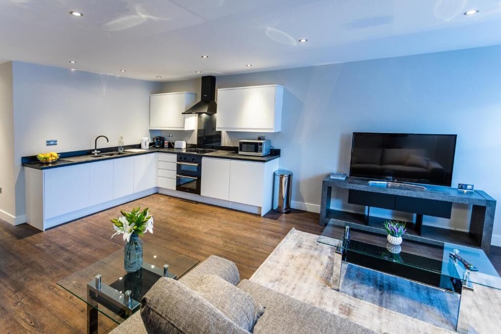 A modern kitchen with white cabinets, a black range hood, and a microwave. There's a dining area with a glass coffee table and a sofa. A flat-screen TV is mounted on a black console table.