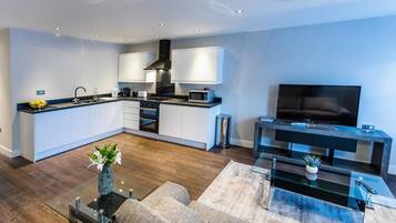 A modern kitchen with white cabinets, a black range hood, and a microwave. There's a dining area with a glass coffee table and a sofa. A flat-screen TV is mounted on a black console table.