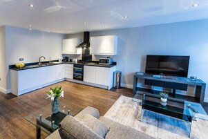 Cocina moderna con armarios blancos, extractor de aire negro y microondas. Hay un comedor con mesa de centro de vidrio y sofá. Un televisor de pantalla plana está montado sobre un mueble consola negro.