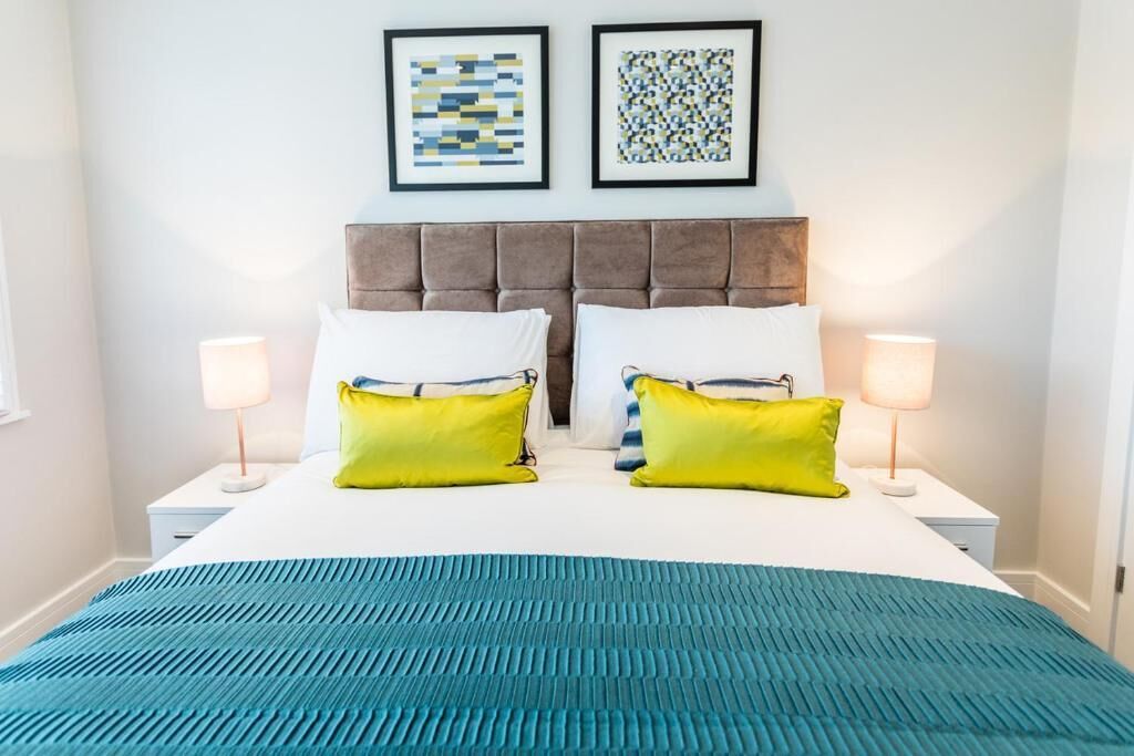 Ein Schlafzimmer mit einem gebürsteten Kopfteil, zwei Nachttischlampen, einer blauen, strukturierten Bettdecke und zwei Kissen mit gelb-weißen Akzenten.