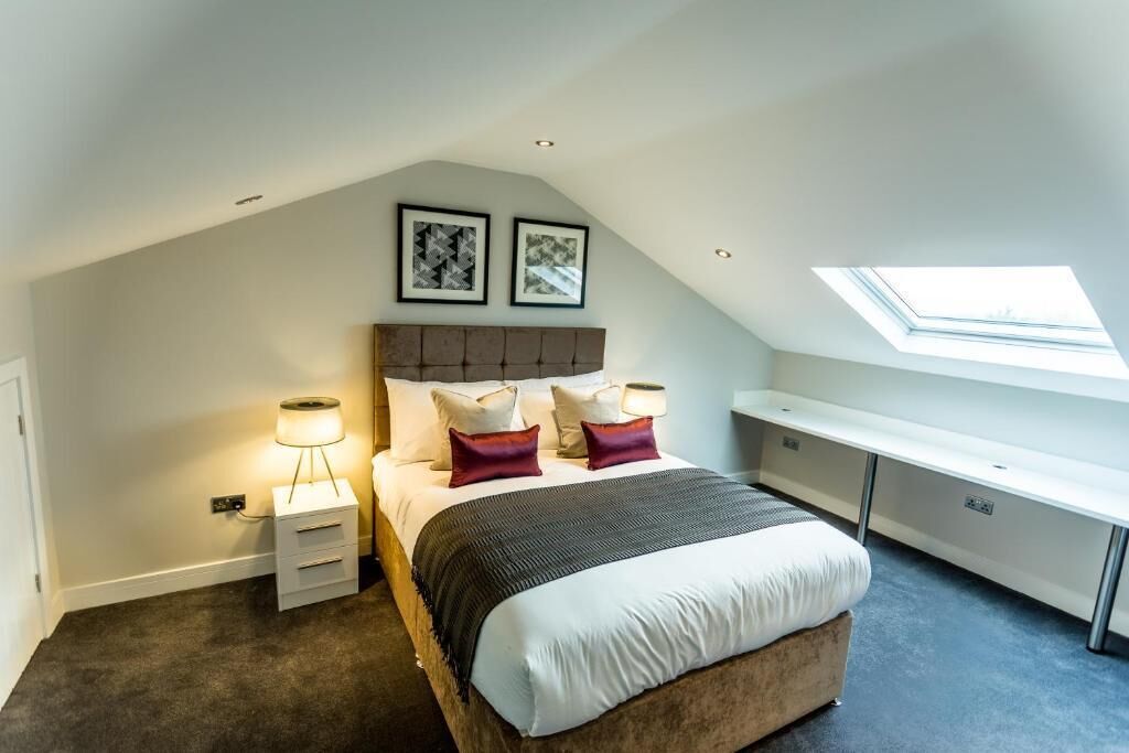 Une chambre avec un plafond en pente, un lit avec du linge de lit blanc et des coussins décoratifs, une table de chevet avec une lampe et un bureau avec une chaise.
