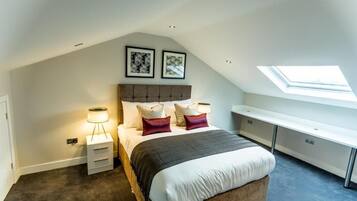 Une chambre avec un plafond en pente, un lit avec du linge de lit blanc et des coussins décoratifs, une table de chevet avec une lampe et un bureau avec une chaise.