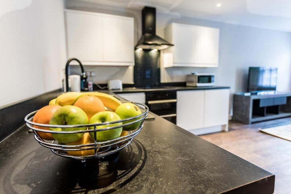 A modern kitchen with a black countertop, a fruit bowl with apples and oranges, white cabinets, and a black range hood.