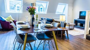 En moderne stue med spiseplads, med et træbord dækket til fire personer, en sofa, et fjernsyn og store vinduer med udsigt.