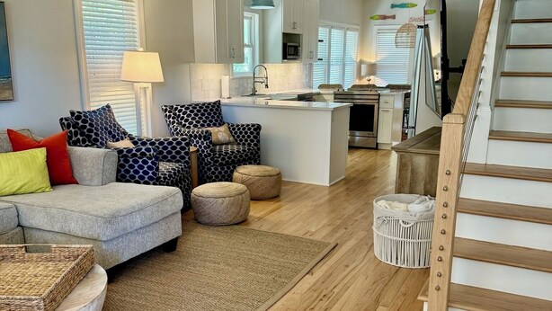 Un salón espacioso con un sofá gris, otomanas y piso de madera. Se ve una cocina al fondo, con una escalera que sube a otro nivel.