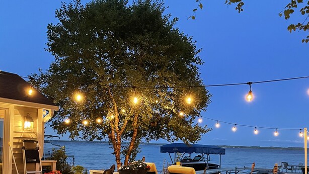 A wooden deck with outdoor furniture, a tree, and string lights against a backdrop of a body of water. - Stunning lake cottage in the heart of wine country (Traverse City)