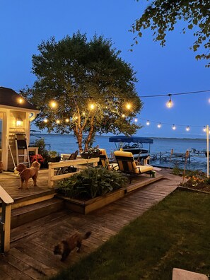 Een houten terras met tuinmeubilair, een boom en lichtjes, met een waterpartij op de achtergrond.
