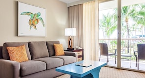 A hotel room with a grey sofa, a blue coffee table, a framed artwork of a sea turtle, a lamp, and a view of palm trees through the balcony doors.