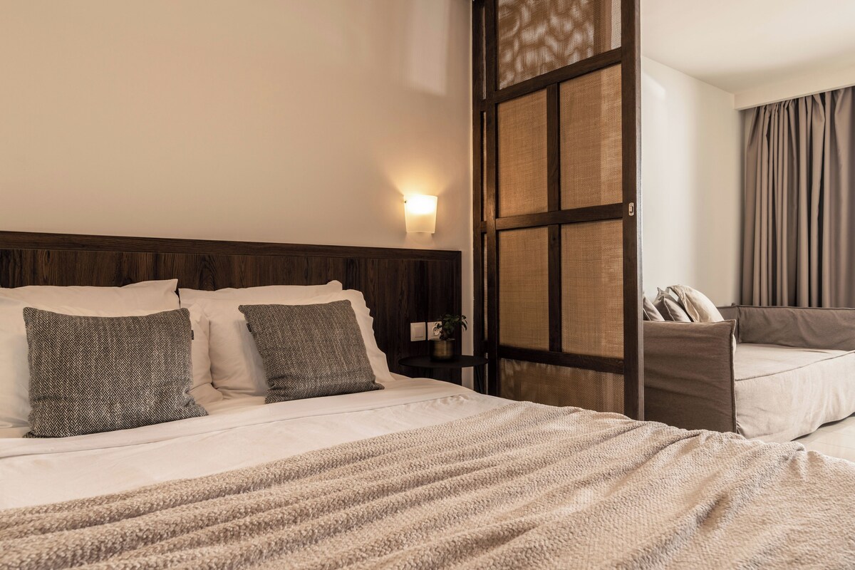 Habitación de hotel con cama, mesita de noche, lámpara de pared y vistas a través de una ventana con cortinas.