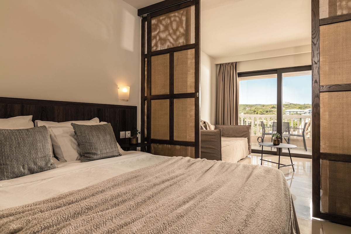 A hotel room with a large bed, a view of the outdoors, and a balcony with a table and chairs.