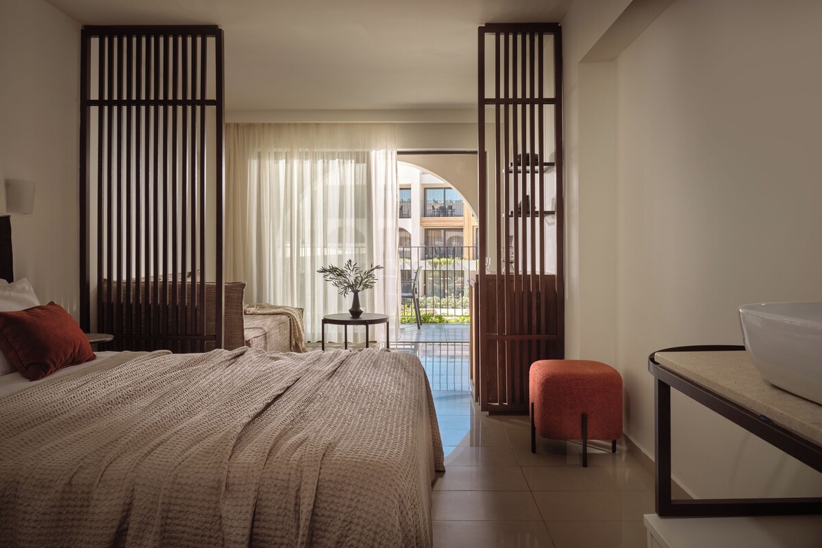 Habitación de hotel con una cama grande, una mesita con una planta, un balcón con vistas a edificios y un escritorio con un cuenco.