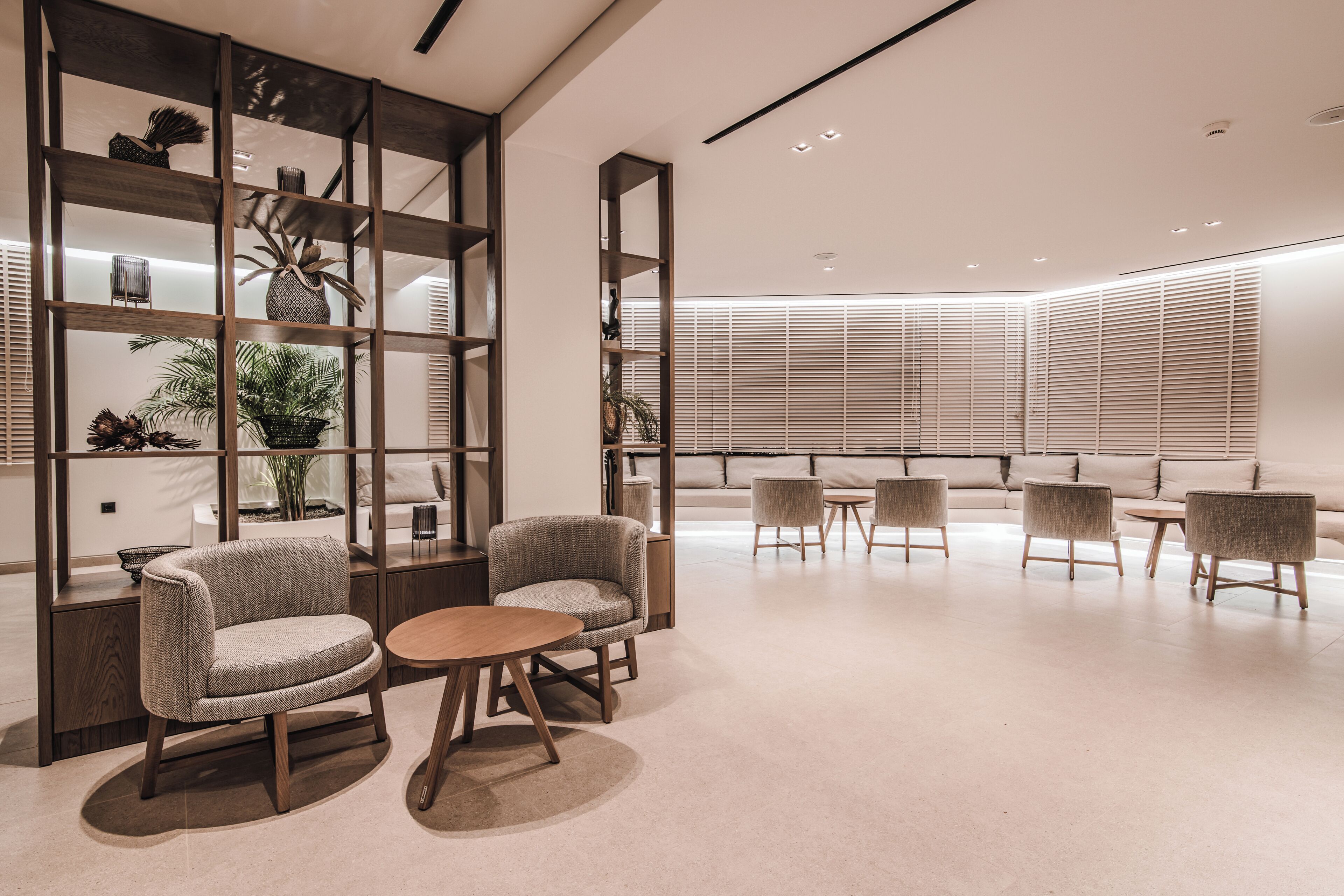 A modern interior with a seating area featuring sofas and armchairs, a wooden bookshelf, and a large window with blinds.