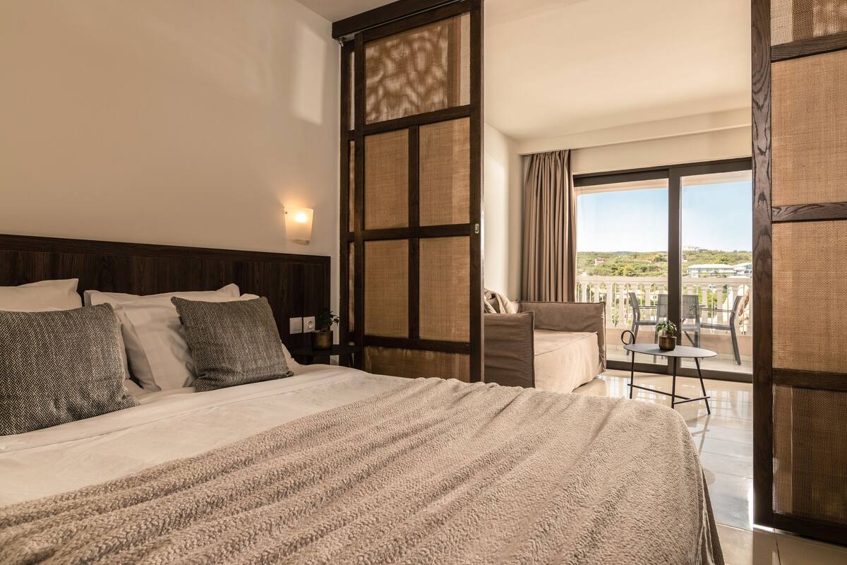 Habitación de hotel con una cama grande, vistas al exterior y un balcón con mesa y sillas.