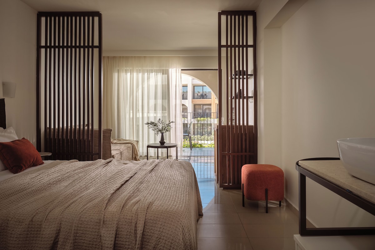 A hotel room with a large bed, a small table with a plant, a balcony with a view of buildings, and a desk with a bowl.