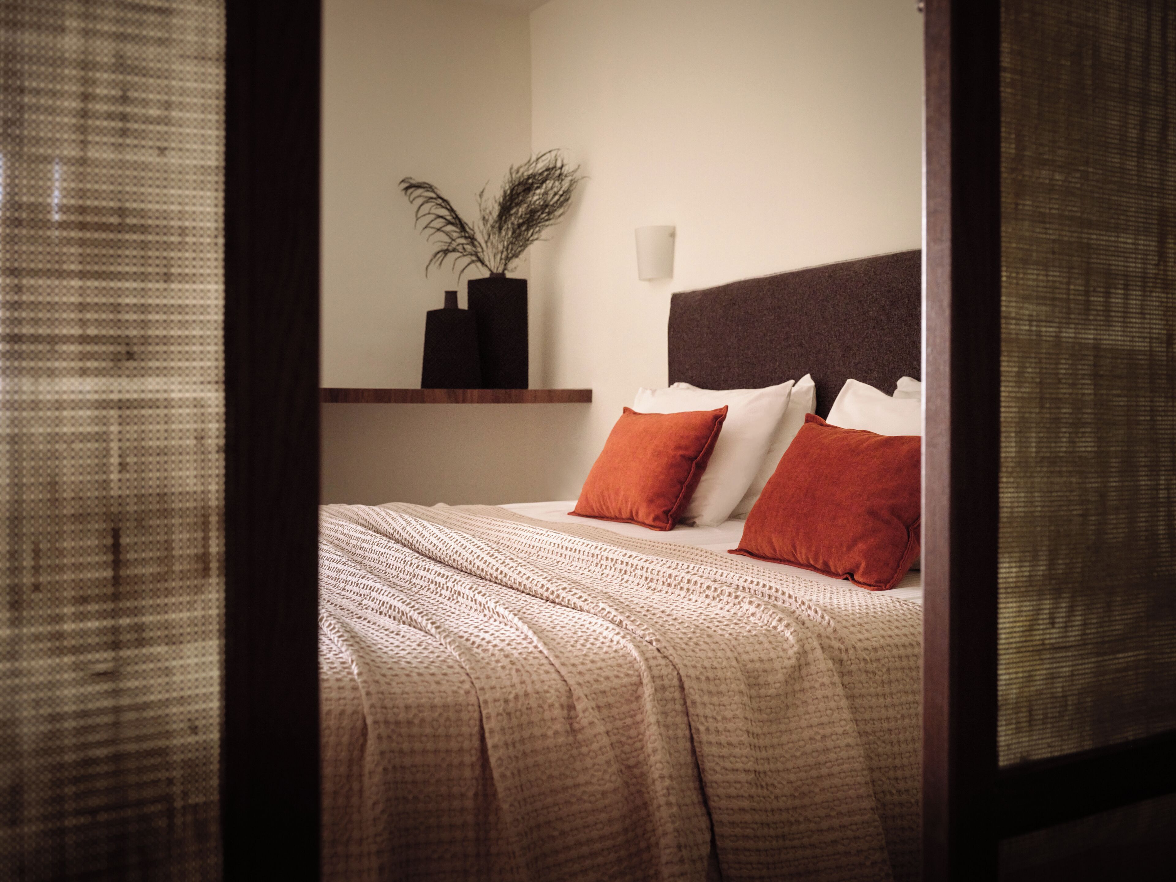 A neatly made bed with a patterned bedspread and two pillows, a small potted plant on a shelf, and a wall-mounted light fixture.