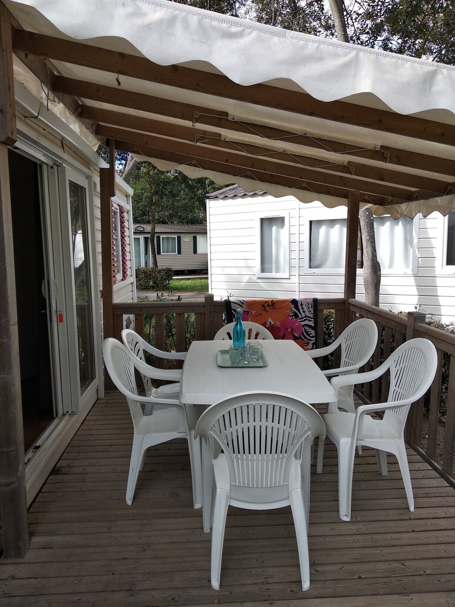 A covered outdoor patio with a white table and chairs, a wooden deck, and a view of a white mobile home in the background.