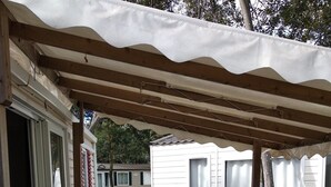 A covered outdoor patio with a white table and chairs, a wooden deck, and a view of a white mobile home in the background. - Your vacation with peace of mind! (Agde)