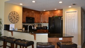 A kitchen with wooden cabinets, a black refrigerator, a microwave, and a clock on the wall. There are three bar stools and a sink. - 3BR/3BA Direct Oceanfront Condo - Magnificent Panoramic View! (daytona beach shores)