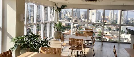 A spacious meeting room with large windows offering a city view, wooden tables and chairs, and potted plants.