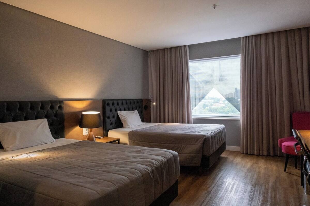 Camera d'albergo con due letti, un comodino con una lampada, una scrivania con una sedia e un'ampia finestra con tende.