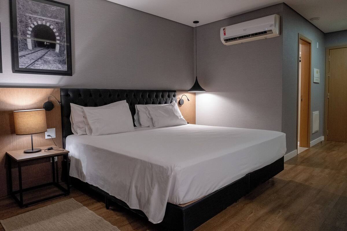 Un letto rifatto con cura in una camera d'albergo, con una testiera, una lampada da comodino e un quadro appeso al muro.