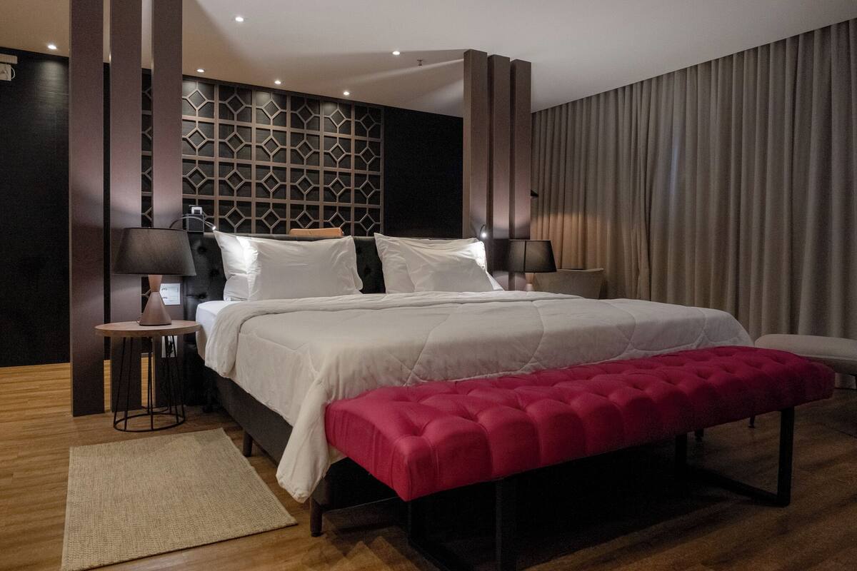 Una moderna camera d'albergo con un letto grande, una panca rossa, due comodini con lampade e un motivo geometrico sulla parete.