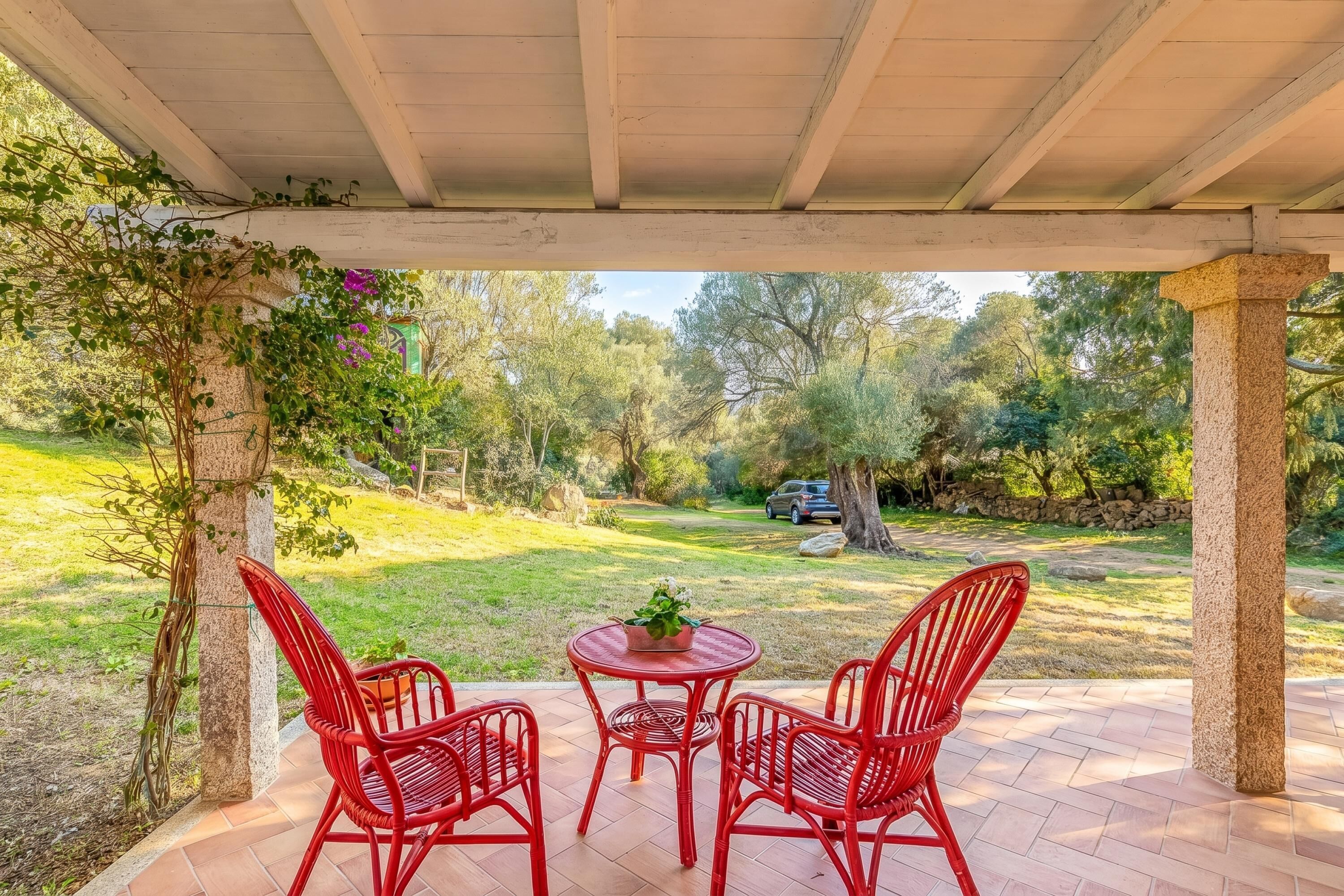 Un patio cubierto con sillas rojas y una mesa, con vistas a un jardín con árboles y un coche al fondo.
