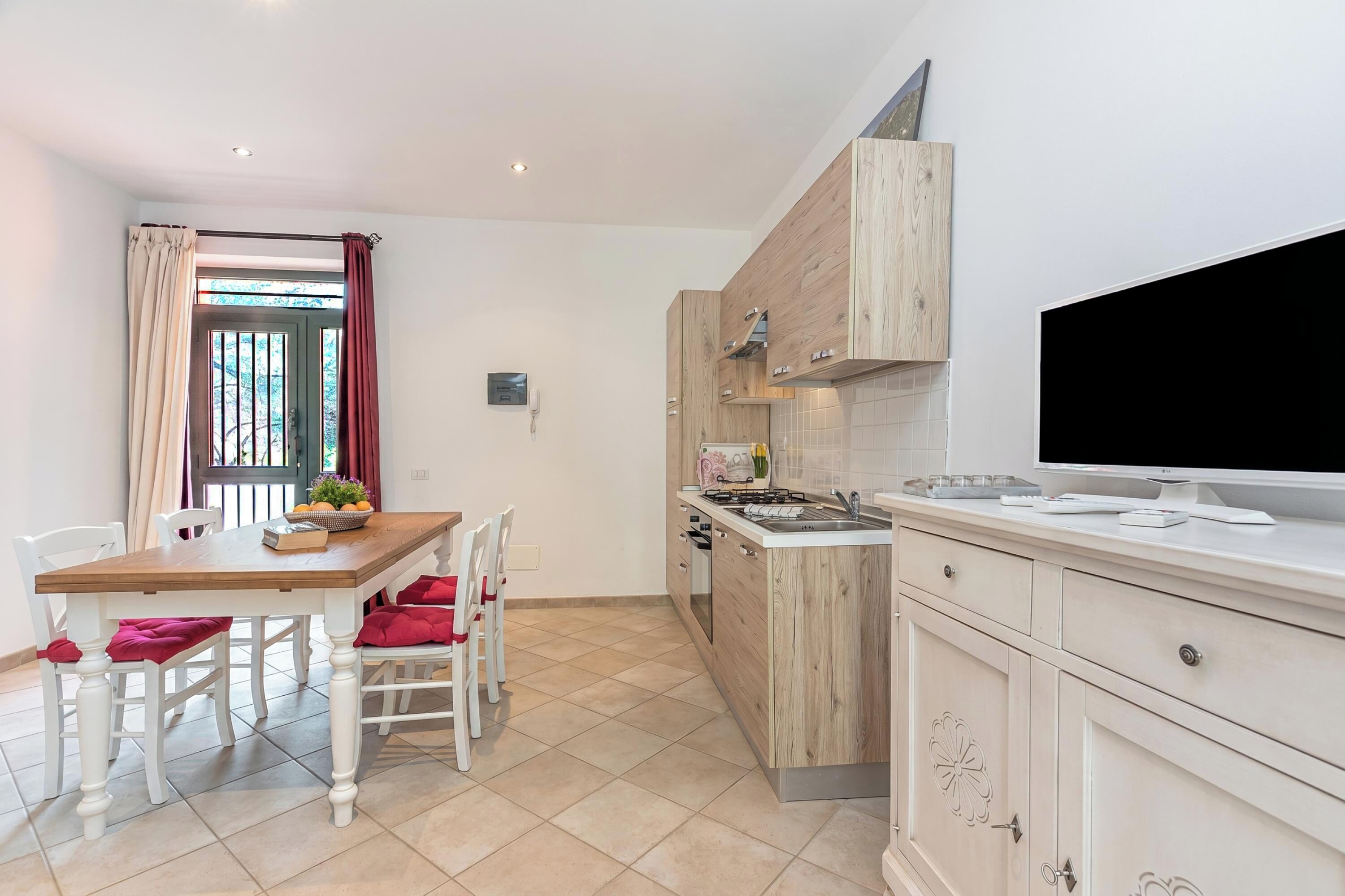 A modern kitchen with a dining area, wooden cabinets, a large TV, and a window with curtains.