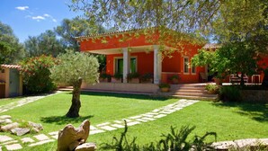 A red building with columns, a stone pathway, and a tree in the foreground. - Domos Pintadas (Olbia)