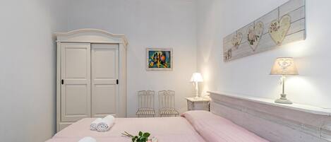A bedroom with a bed, a wardrobe, a nightstand with a lamp, two chairs, and a wall art piece.