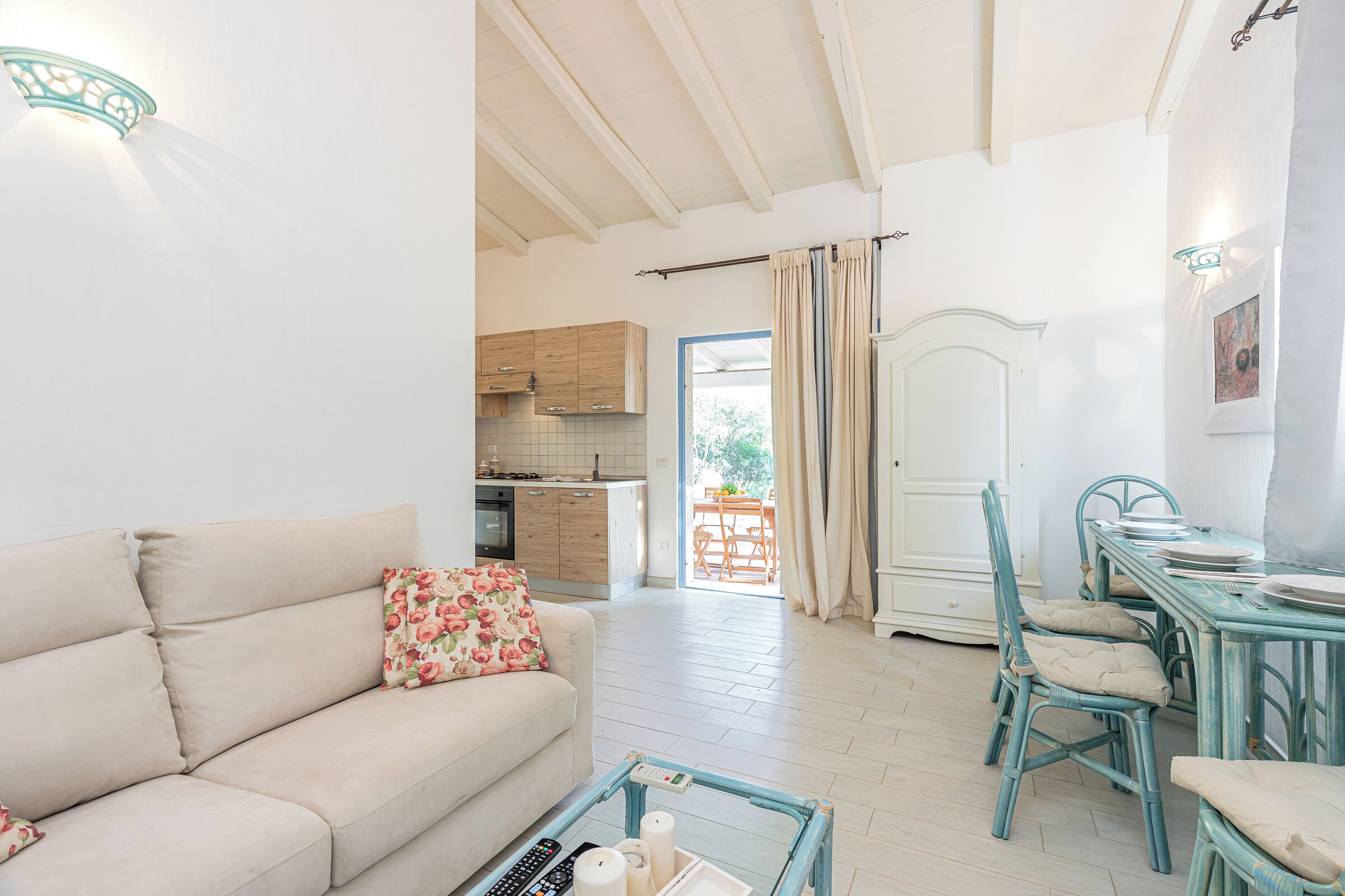 A living room with a sofa, a coffee table, a dining table with chairs, a kitchen area, and a door leading outside.