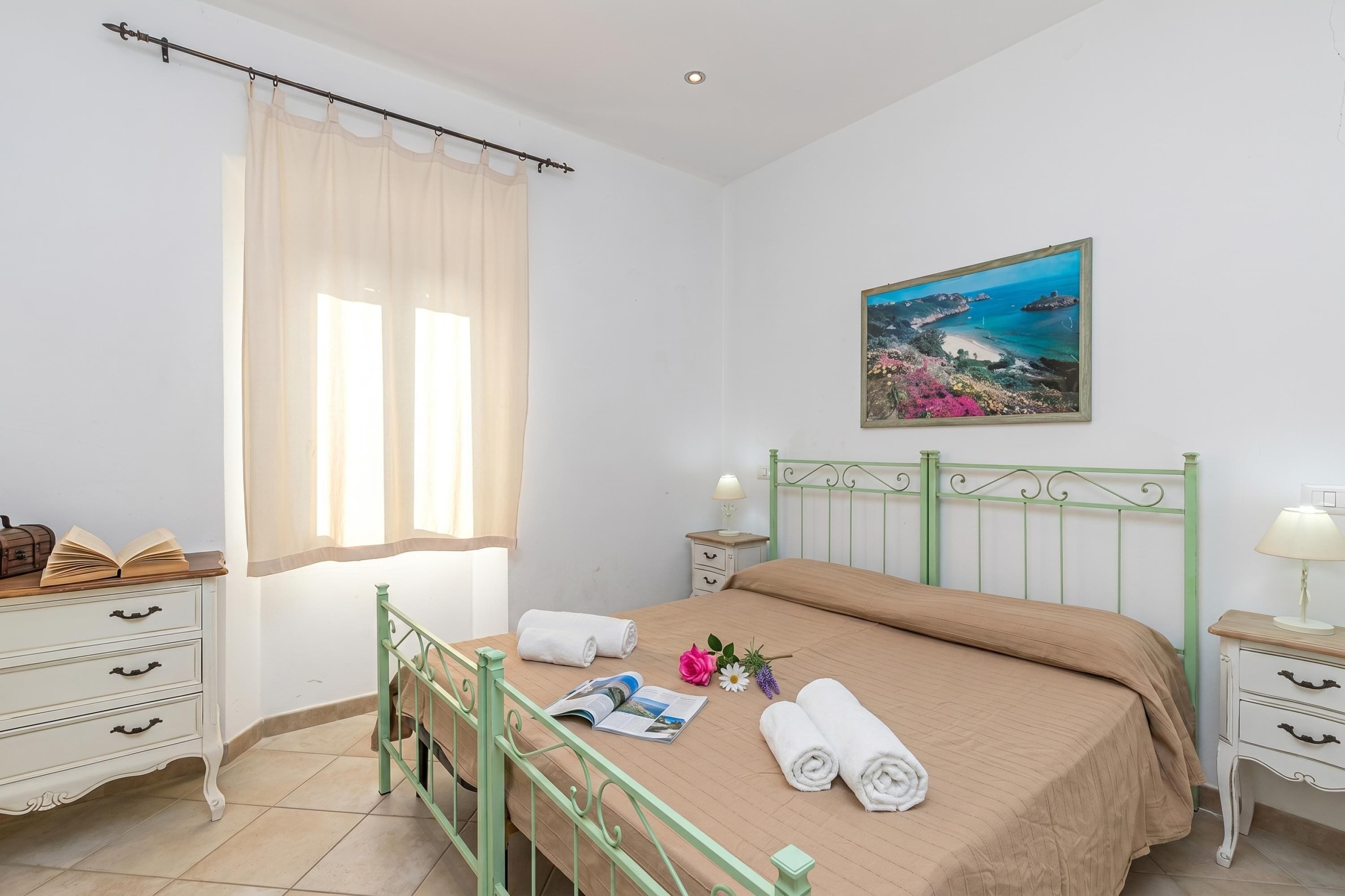 Un dormitorio con una cama, una mesita de noche con lámpara, una ventana con cortinas traslúcidas, un cajón junto a la cama y un cuadro en la pared.