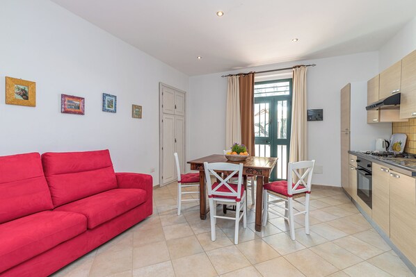 A modern living room with a red sofa, a wooden dining table with four chairs, and a kitchen area with wooden cabinets and a stove. - Domos Pintadas (Olbia)