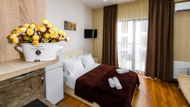 Quarto de hotel com cama, televisão, janela com cortinas, pequena cozinha e um vaso de flores numa prateleira.