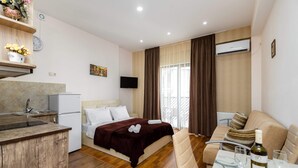 Quarto de hotel com cama, sofá, mesa de jantar com garrafa e copos, cozinha e uma grande janela com cortinas.