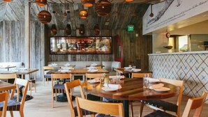 Un restaurant amb taules i caires de fusta, llums penjants de coure i una zona de bar al fons.