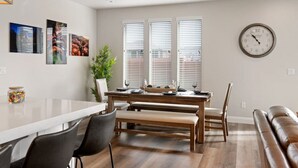 A modern dining area with a wooden table, chairs, and a bench. There are three windows with blinds, a clock on the wall, and a plant on the table. - Large 5 Bed 5 Bath Home w/Private Hot Tub (Washington)