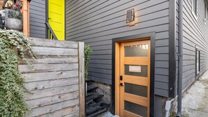 Puerta de madera con un panel de vidrio y un letrero de bienvenida, frente a la fachada gris de una casa.
