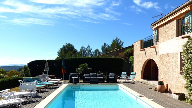 Une piscine entourée d’une terrasse en bois et d’un bâtiment à la façade en pierre, avec des chaises longues et des parasols à proximité.