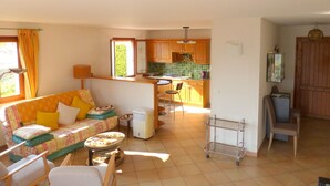 Un salon chaleureux avec un canapé, des fauteuils et une petite table. On aperçoit, au fond, une cuisine avec des meubles en bois et un plan de travail recouvert de carreaux.