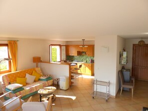 A cozy living room with a sofa, chairs, and a small table. A kitchen with wooden cabinets and a tiled backsplash is visible in the background.