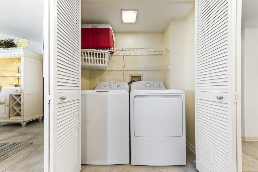 A laundry room with two white washing machines and a red basket on a shelf.