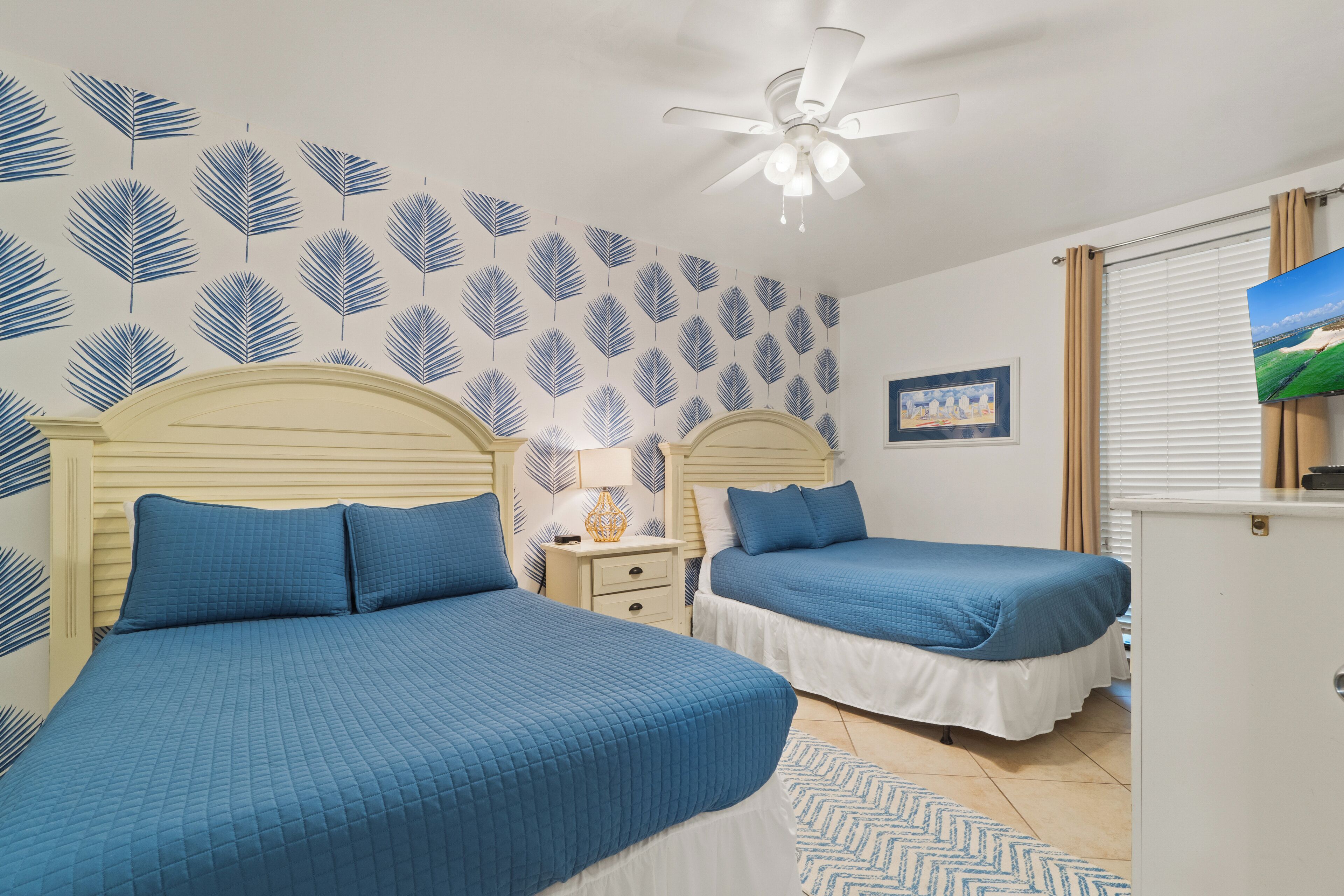 Un dormitorio con dos camas, un ventilador de techo, una mesita de noche y un cuadro en la pared.